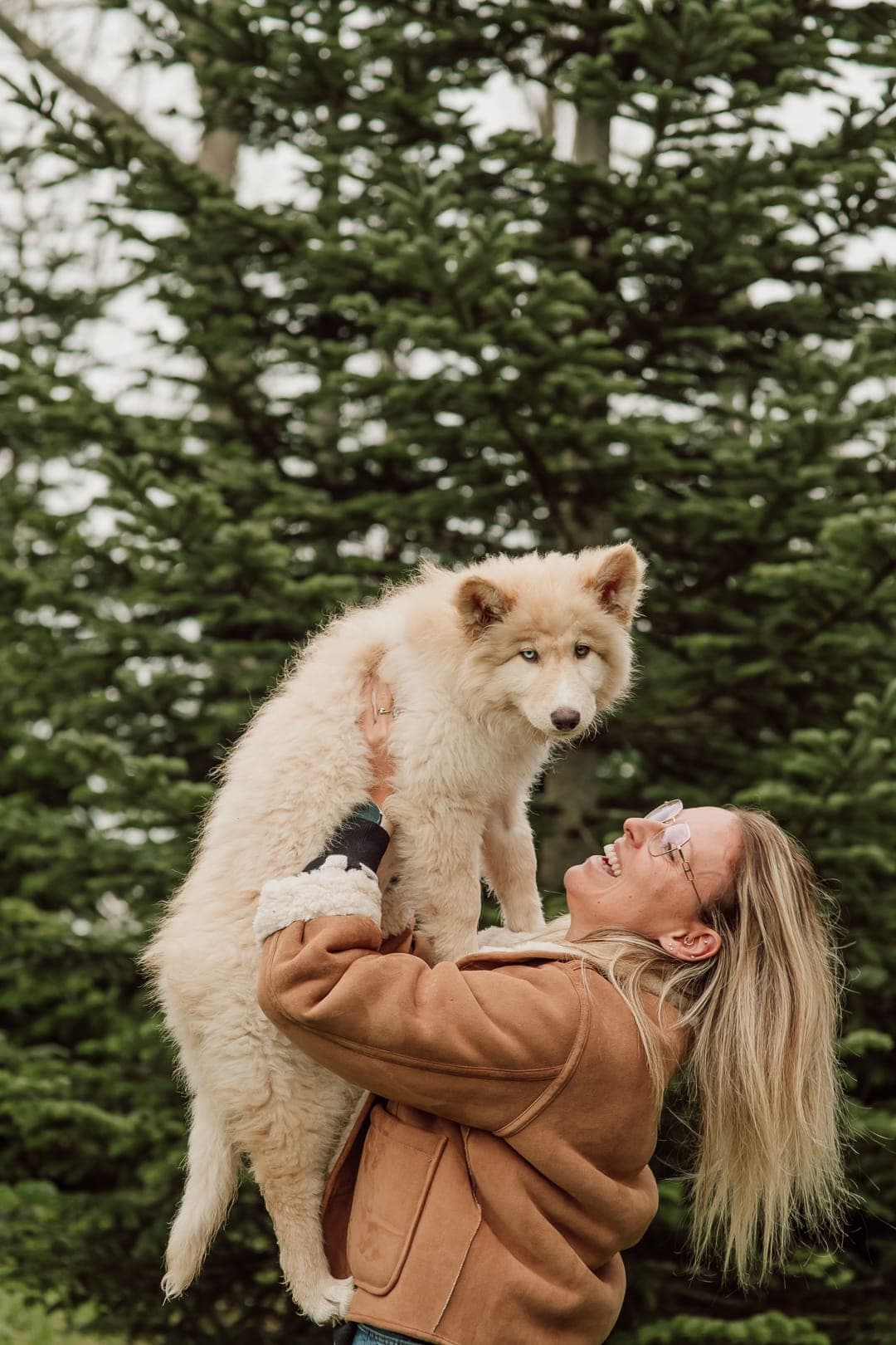 Stéphanie tilt lachend een lichte pup op, met dennenbomen op de achtergrond
