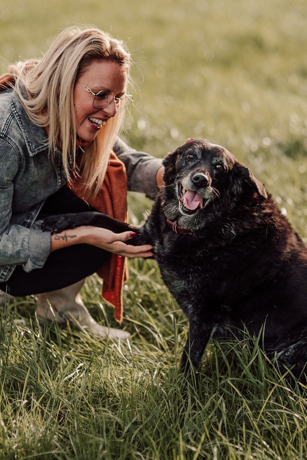Stéphanie zit gehurkt in het gras bij een oudere zwarte hond die haar hand besnuffelt